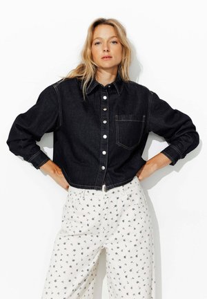 Veste en denim foncé, cropped, avec un col, dotée d'une poche poitrine et de boutons argentés, associée à un pantalon blanc à motif.