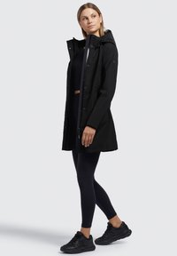 Zwarte waterdichte jas met een capuchon, voorzien van een ritssluiting en knoopsluiting, en zijzakken, gedragen over een donkere top en leggings.