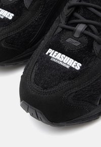 Črne teniske z mehkimi teksturami, iz semiša in z gumijastim podplatom. Ponašajo se s poudarjenim belim logotipom "PLEASURES DMX RIDE" na strani.