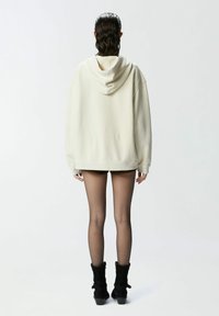 Felpa oversize color crema con cappuccio e cordino, texture morbida e polsini a costine, abbinata a pantaloni corti scuri e stivali neri alla caviglia.