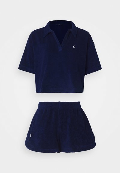 Σέτ lounge από χνουδωτό ύφασμα σε ναυτικό μπλε, περιλαμβάνει κοντό πόλο μπλουζάκι με γιακά και κοντές σορτσάκια, και τα δύο με μικρές λογότυπες λεπτομέρειες.