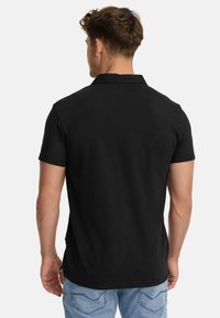 Camiseta polo de algodón negro con cuello y mangas cortas. Presenta un dobladillo recto y una textura sutil, combinada con jeans de color azul claro.