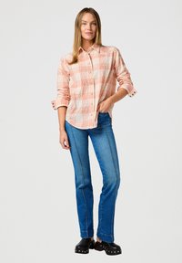 Camicia a quadri in tessuto arancione chiaro e bianco, con colletto a bottoni e maniche arrotolate. Abbinata a jeans blu di denim e zoccoli neri con borchie.