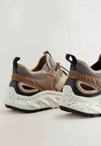 Baskets sportives avec un schéma de couleurs gris, marron et beige, présentant des surfaces texturées, une semelle blanche mouchetée et des languettes pour un enfilage facile.