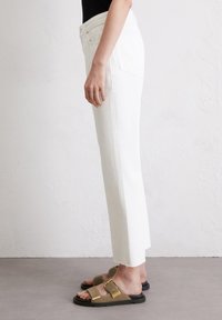 Witte rechte jeans met een rafelige zoom, gecombineerd met bruine slidesandalen met gouden gespen. Achtergrond is neutraal en gestructureerd.