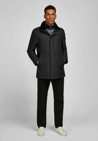 Zwarte geïsoleerde jas met een nepbontkraag, ritssluiting aan de voorkant en drukknoopsluitingen. Voorzien van zijzakken en een rechte snit, gedragen over zwarte broek.