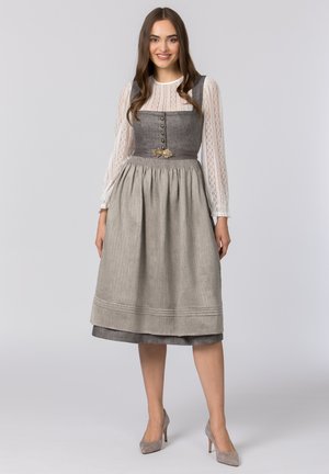 Junge Frau mit langen braunen Haaren, die ein graues traditionelles Kleid mit knöpfbarem Mieder, beige Schürze, weiße Spitzenbluse und graue Absätze trägt und lächelt.