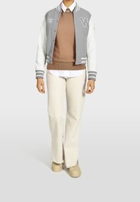 Gray and white varsity jacket with embroidered lettering, layered over a brown sweater and white shirt, paired with beige wide-leg pants.