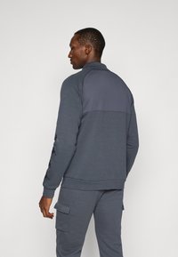 Sudadera de cremallera azul marino con un panel superior texturizado, adornada con acentos bordados en las mangas y puños acanalados, combinada con pantalones a juego.