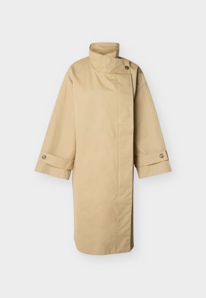 Bēša gara trenčmētelis ar augstu apkakli, asimetrisku priekšpuses pogu aizdari un pogām dekorētiem aproču galiem uz vienkārša gaiša fona.