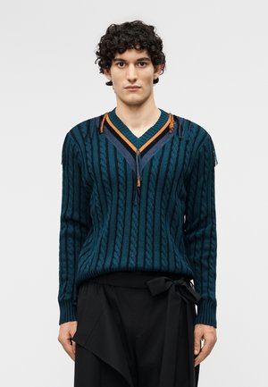 Jeune personne aux cheveux bouclés et foncés portant un pull en tricot torsadé couleur sarcelle avec un col en V rayé et un pantalon noir ample attaché à la taille.