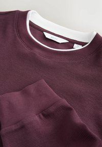 Bordeaux Sweatshirt aus strukturiertem Stoff, mit weißem Rundhalsausschnitt und gerippten Bündchen. Mit sichtbarem Markenschild und einer gestalterischen Taschenkontur.