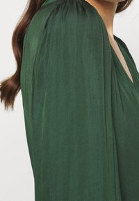 Blusa verde con un design fluido, tessuto testurizzato e maniche a sbuffo plissettate. Superficie liscia con scollatura a V.