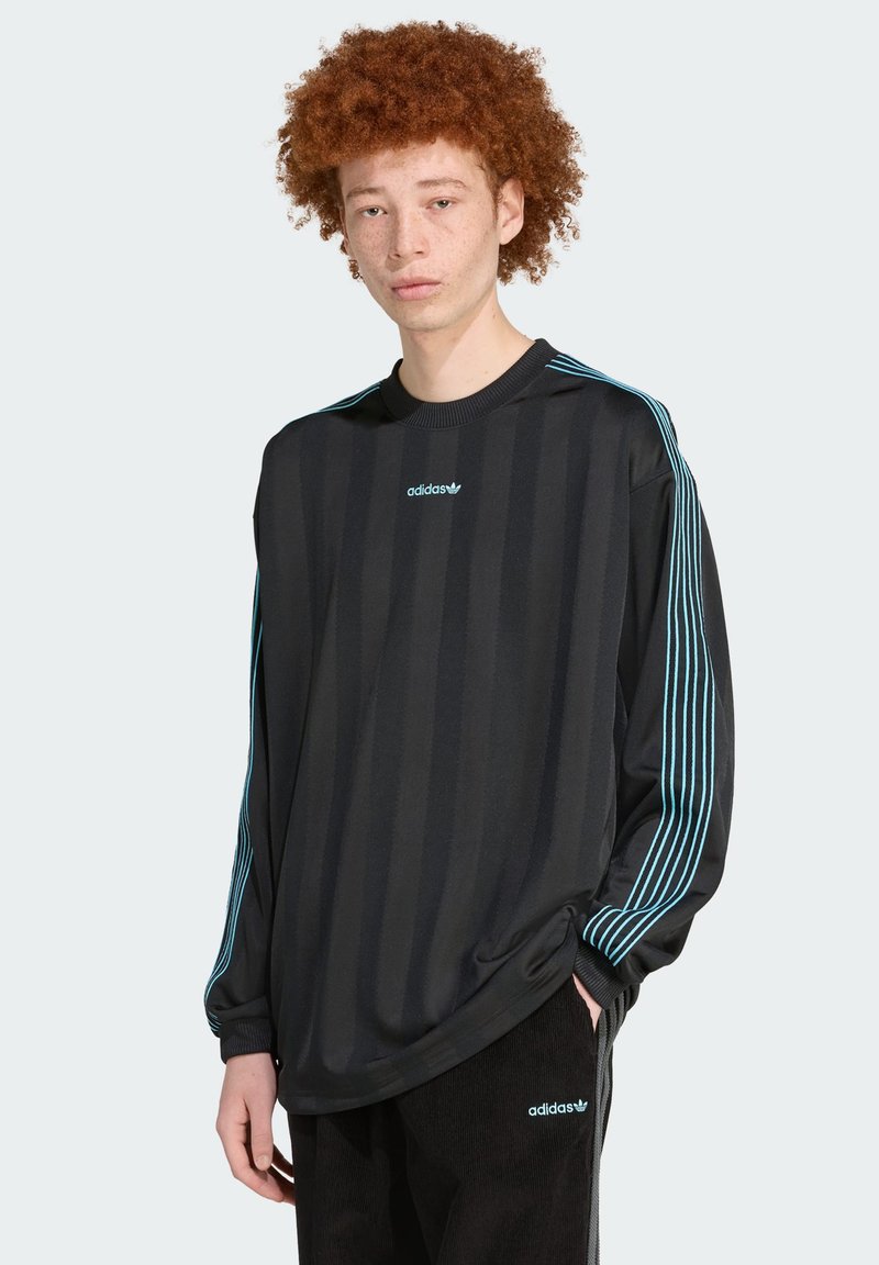 Μαύρο μπλουζάκι με μακριά μανίκια και κατακόρυφες ρίγες, με ανοιχτό μπλε λωρίδες στο πλάι και λογότυπο στο στήθος. Ούτε το ύφασμα φαίνεται λείο και αθλητικό.