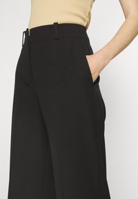 Pantalon noir sur mesure à taille haute, avec poches latérales et texture lisse ; doté d'une fermeture éclair et d'un design minimaliste.