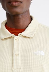 Beige vaffelstrikket polo med krave, der har to cremefarvede knapper og et hvidt logo på venstre bryst. Underlaget er orange.