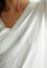 Bílý top s výstřihem do V a vlnitým lemem, vyrobený z jemného materiálu, s jednoduchým designem a hladkým povrchem.