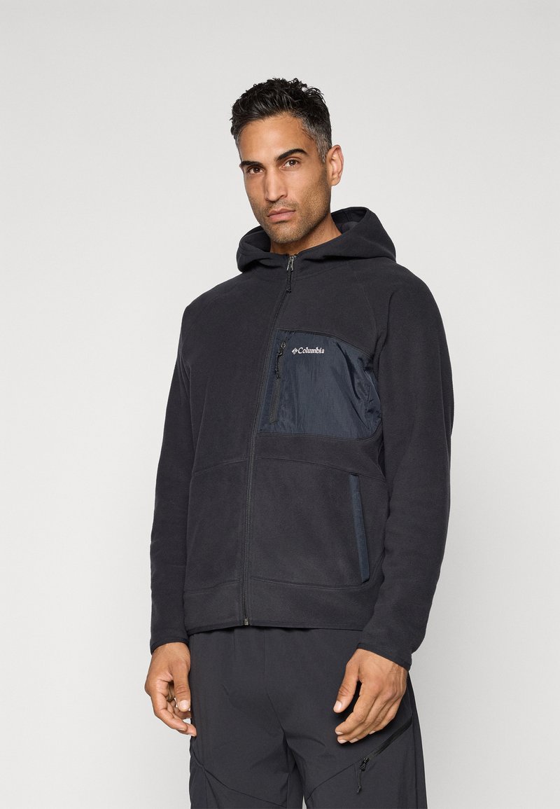 Mann iført en svart hettejakke med glidelås og fleece, med Columbia-logo på brystet, stående mot en ensfarget lys bakgrunn.