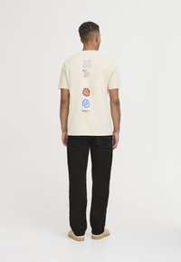 Camiseta de algodón color crema con gráficos coloridos en la parte posterior: espiral morada, engranaje rojo, remolino azul y el texto "NO IMPORTA QUÉ". Llevada con pantalones negros.