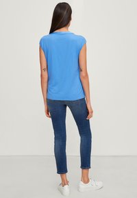 Blaues kurzärmliges Oberteil mit rundem Hals, kombiniert mit dunklen engen Jeans und weißen Sneakers. Der Stoff wirkt glatt mit einer einfarbigen Textur.