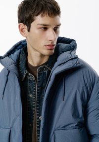 Blå puffjacka med huva, som har en slät yta och dragkedjestängning. Lager på en mörk jeansjacka med knappdetaljer.