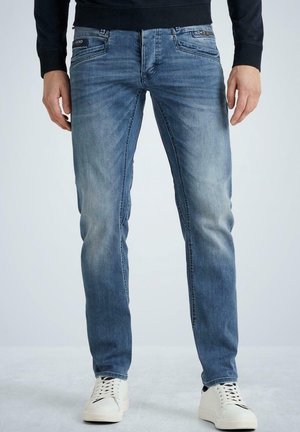 Man wearing faded blue jeans and white sneakers, standing with hands relaxed at sides against a plain background.