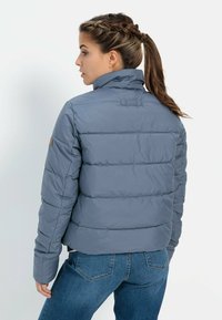Grijze pufferjack met een hoge kraag, voorzien van horizontale gestikte details en een aansluitende pasvorm, gecombineerd met blauwe denim jeans.