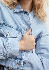 Giacca in denim blu chiaro con chiusura a bottoni, cuciture visibili e vestibilità rilassata. Presenta una tasca sul petto e maniche arrotolate.