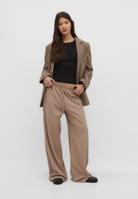Kvinde i løse beige bukser, sort top og ternet blazer står mod en ensfarvet baggrund og justerer sit livrem.