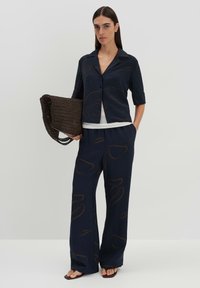Ensemble deux pièces marine comprenant une chemise texturée à boutons et manches retroussées ainsi qu'un pantalon large avec des motifs abstraits marron. Modèle tenant un sac en osier marron.