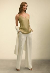 Gouden satijnen cami met dunne bandjes, gecombineerd met witte wijde broekspijpen. Een lichtbeige sjaal hangt losjes over één schouder, sandalen met bandjes.