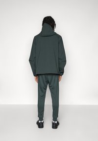 Grønn, lett jakke med hette, paret med matchende bukser. Stoffet har en glatt tekstur og en avslappet passform. Svarte joggesko fullfører antrekket.