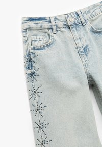 Lysblå denim jeans med en falmet finish, der har et dekorativt sort rhinestonesnefnugmønster langs siden.
