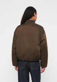 Jachetă bomber căptușită maro, cu guler striat și tiv elastic. Textură netedă, croială ușor largă. Purtați cu pantaloni din denim albastru.