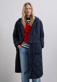 Navy quilted long coat with a hood, featuring a front pocket and snap closures. Worn over a red sweater and striped top with blue jeans.