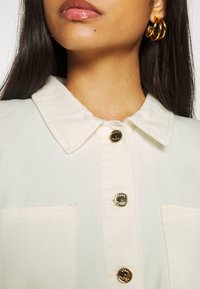 Chaqueta de mezclilla crema con cuello puntiagudo, cierres de botón dorados y dos bolsillos frontales con costuras visibles.