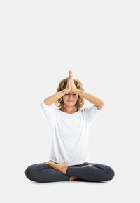 Witte lange mouw top, donkere legging, en een zittende figuur met handen in gebedshouding en gesloten ogen op een eenvoudige achtergrond.