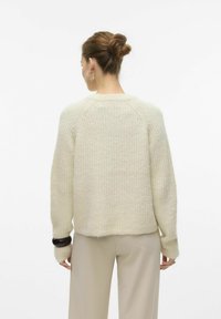 Pull en tricot beige clair avec une coupe décontractée, texture côtelée et manches raglan ; il présente un ourlet arrondi et des accents de poignets foncés contrastants.