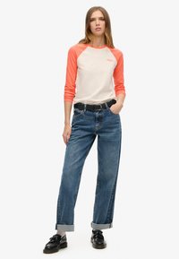 Crème shirt met raglanmouwen en oranje mouwen, gecombineerd met losse blauwe jeans met opgerolde manchetten en zwarte schoenen met kwastjes.