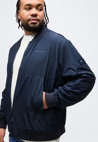 Άνδρας με πλεγμένα μαλλιά, φορώντας ναυτικό μπουφάν Tommy Hilfiger με τα χέρια στις τσέπες, κοιτάζοντας προς τα αριστερά του, πάνω σε ένα απλό φωτεινό φόντο.