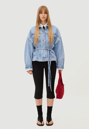 Femme aux cheveux longs et raides portant une veste en denim bleu clair ceinturée, un pantalon noir court, des chaussettes noires avec des sandales, tenant un sac rouge.