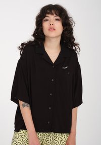Chemise noire à manches courtes avec col, dotée d'une poche et d'un devant boutonné. Tissu doux avec une coupe décontractée, portée sur un short jaune à motifs.