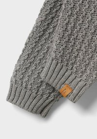 Manica di un maglione grigio lavorato a maglia con un motivo testurizzato e polsino a coste. Un piccolo etichetta in pelle è visibile vicino al polsino.