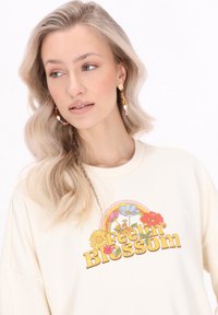Felpa color crema con grafica "Feelin' Blossom" che presenta fiori e un arcobaleno. Il modello indossa orecchini colorati di perline.
