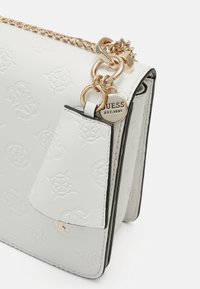 Hvid faux læder håndtaske med præget logo, guld kædetras, og detaljeret hardware. Har en sidelomme og sort kant.