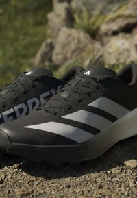 Μαύρα και άσπρα παπούτσια Adidas Terrex για τρέξιμο σε μονοπάτι πάνω σε χαλίκι με φυσικό φόντο από πέτρες και γρασίδι.