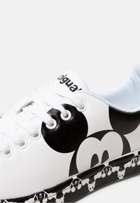 Baskets blanches avec un visage de Mickey Mouse noir sur le côté et un motif Mickey sur la semelle noire, avec des lacets blancs et le nom de la marque sur la languette.