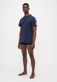 Marineblå t-skjorte med korte ermer, har trykket hvit merkevare logo på skuldrene, laget av myk bomull. Kombinert med svarte shorts.