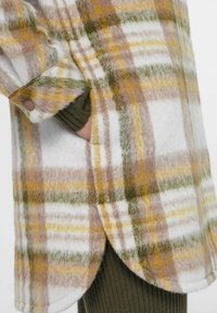 Geruite jas van zacht, fuzzy materiaal, met gele, groene en bruine patronen, een afgeronde zoom en zijzakken. Textuur van de stof is duidelijk zichtbaar.