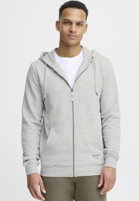 Siva zip-up dukserica s kapuljačom na vezanje, prednjim džepovima i rebrastim manžetama. Ima suptilnu oznaku na donjem rubu.